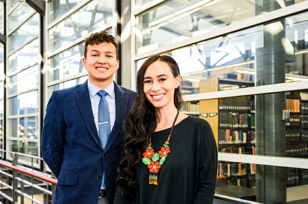 Portrait of Lorenzo Gudino and Catelin Aiwohi standing in the atrium of the law school with book shelves in background