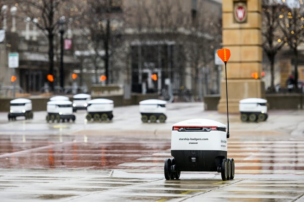 Several food robots roll along a rainy walkway.