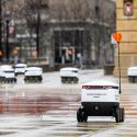 Several food robots roll along a rainy walkway.