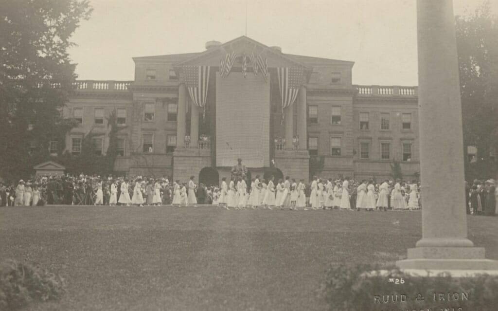 A large group of women in white dresses standing in front of Bascom Hall with a large banner and American flags