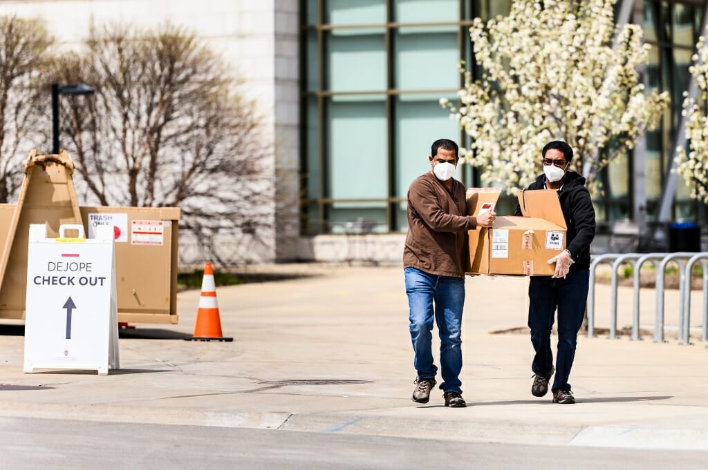 Two men walk down a campus sidewalk carrying things.