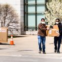 Two men walk down a campus sidewalk carrying things.