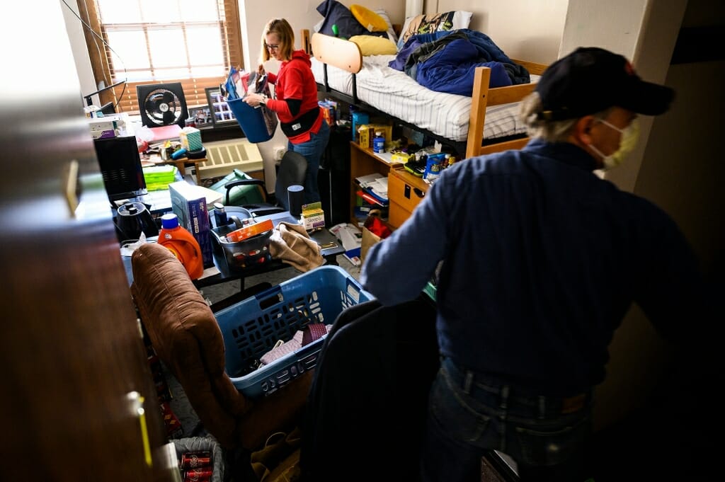 Two people pick up things in a crowded residence hall room.