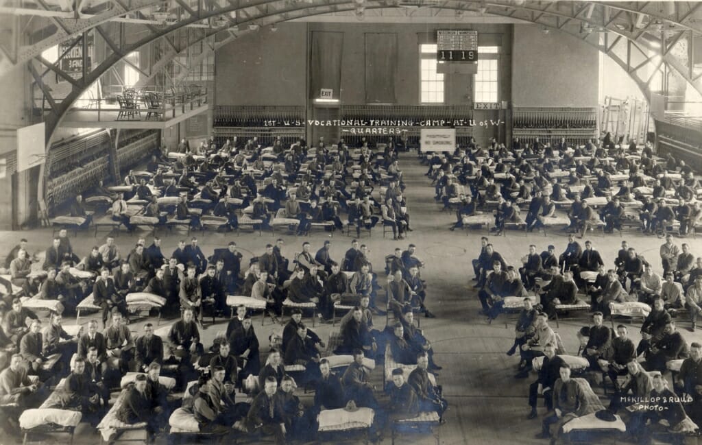 Dozens of cots with men in military uniforms sitting on them in large room with arched ceiling beams