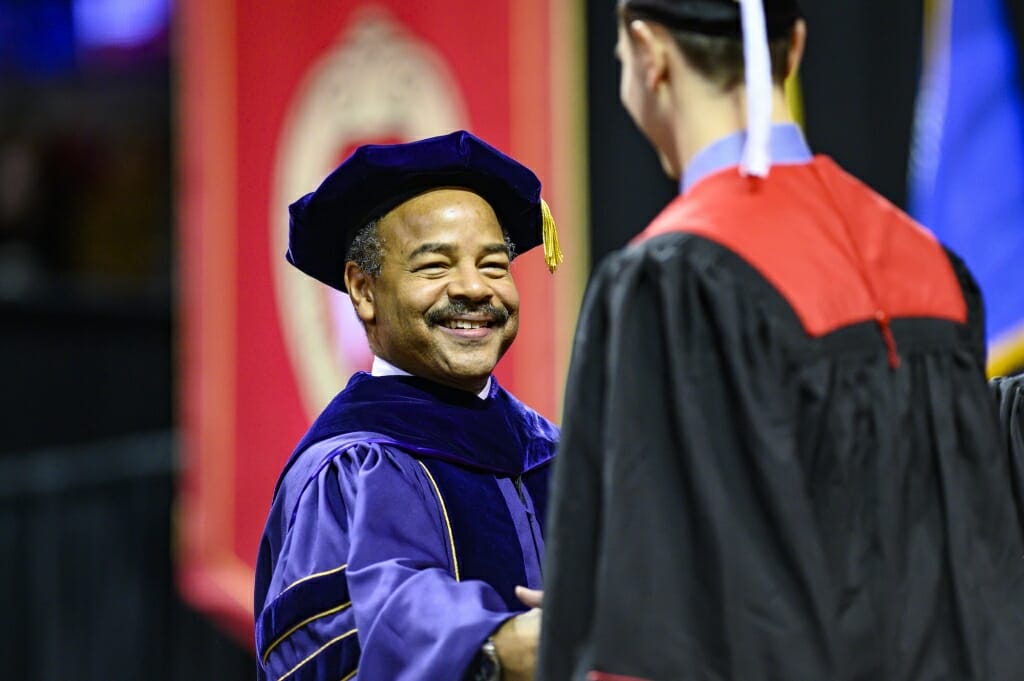 Eric Wilcots in academic regalia shaking hands with a graduate in front of a red W banner