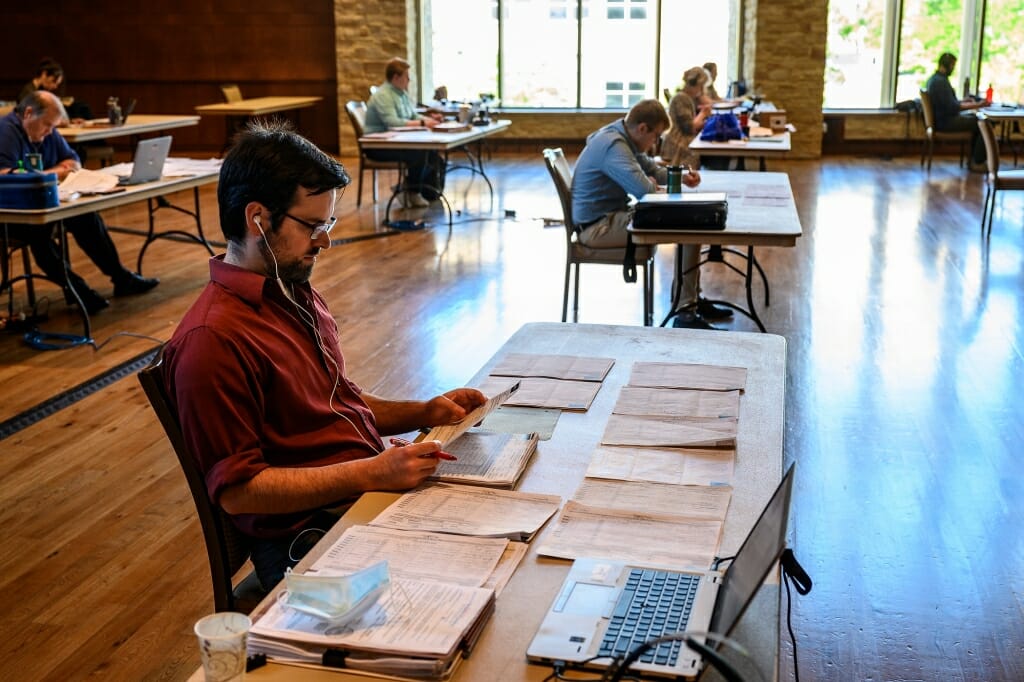 Office workers are seated at desks separated by the required 6 feet.