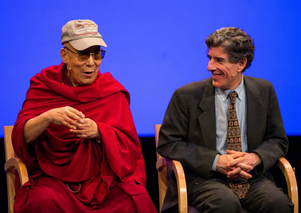 The Dalai Lama wearing a red robe sits next to Richard Davidson who is wearing a suit.