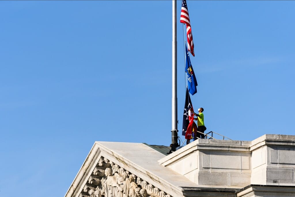 A worker standing on the roof of the State Capitol raises the Juneteenth flag along with the American and state flags.