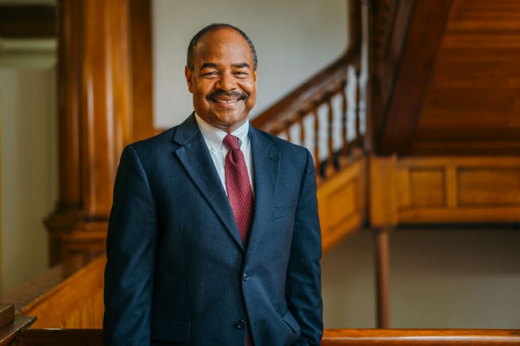 A photo portrait of Eric Wilots. Wilcots is standing in a wood-paneled foyer. Wilcots is wearing a dark suit with a white shirt and a red tie. He has short, dark hair and a mustache. He is smiling to the camera.