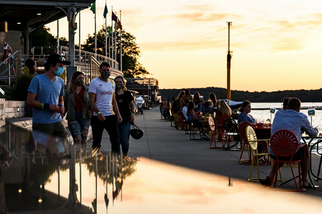 A terrace sunset is reflected off a table; people walk by with masks.