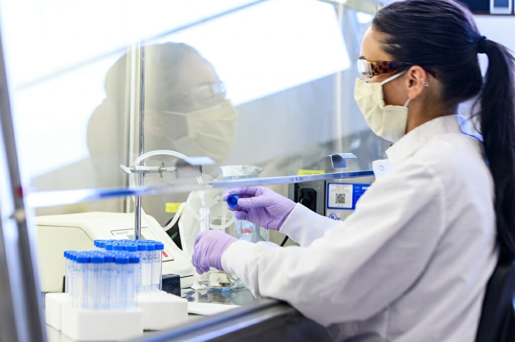 A woman wearing a white lab coat, gloves and face mask works with test tubes.