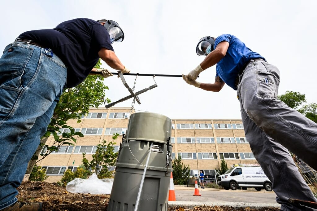 Two plumbers lifting a large cylinder out of a maintenance hole in front of a residence hall