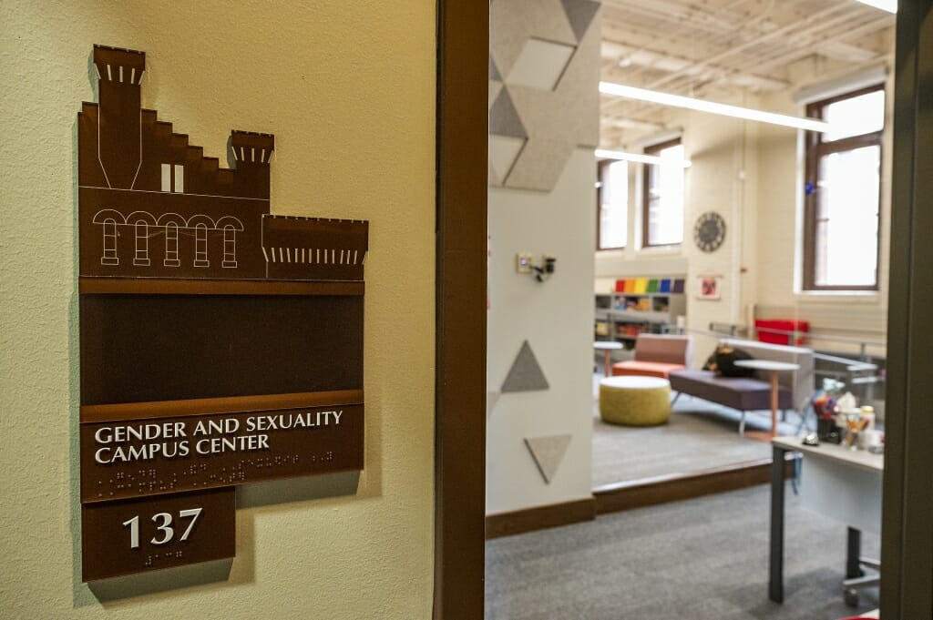 Wall sign for Gender and Sexuality Campus Center outside doorway looking in at room with chairs and shelves