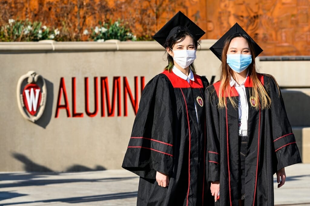 2 people in caps, gowns and face coverings standing in front of the word 