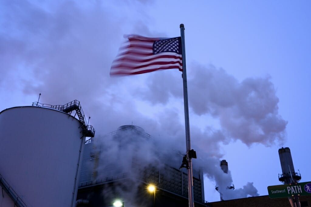 An American flag flutters in the foreground and steam rises from the plant.