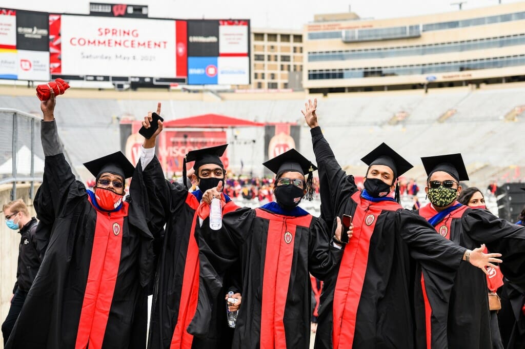 A group of five graduates celebrate, flinging their arms wide.