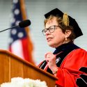 Blank in academic regalia speaking at a podium in front of a U.S. flag