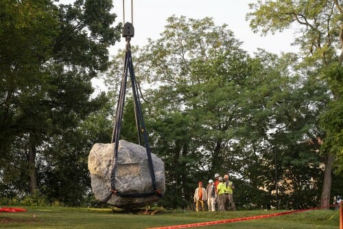 Hanging from long straps attached to a crane, the rock hovers a couple feet above the ground.