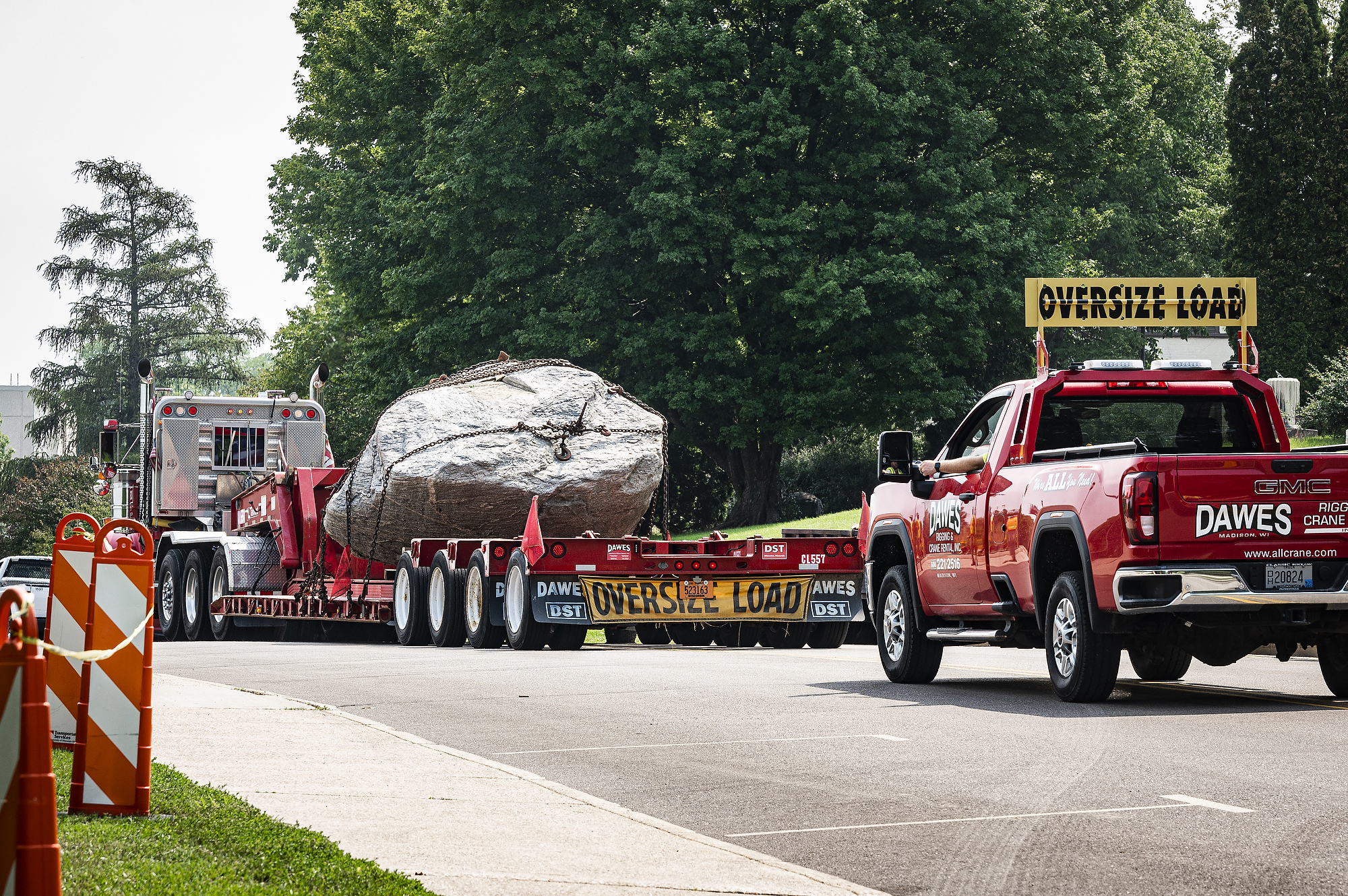 A flatbed truck drives down the road with the rock in its bed, secured by chains. A pickup truck with the sign "oversize load" follows.