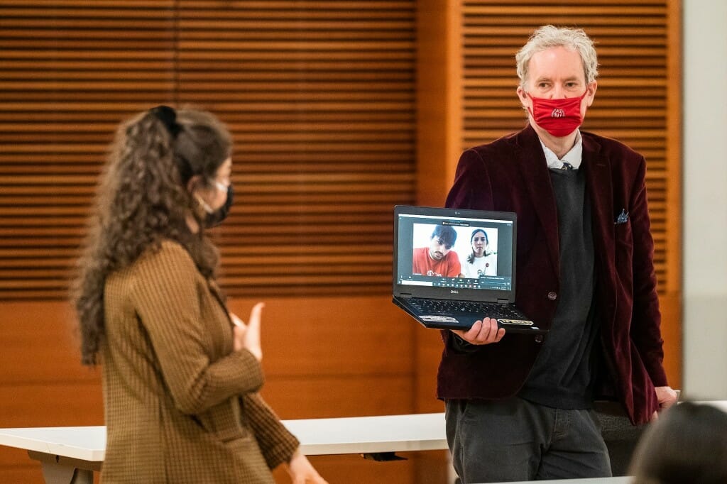 A professor holds a laptop with 2 students on the screen while another student watches in person