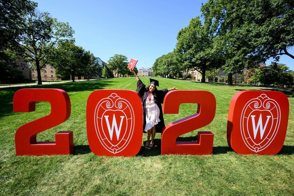 A person in cap and gown standing next to oversized numbers 2020 on Bascom Hill