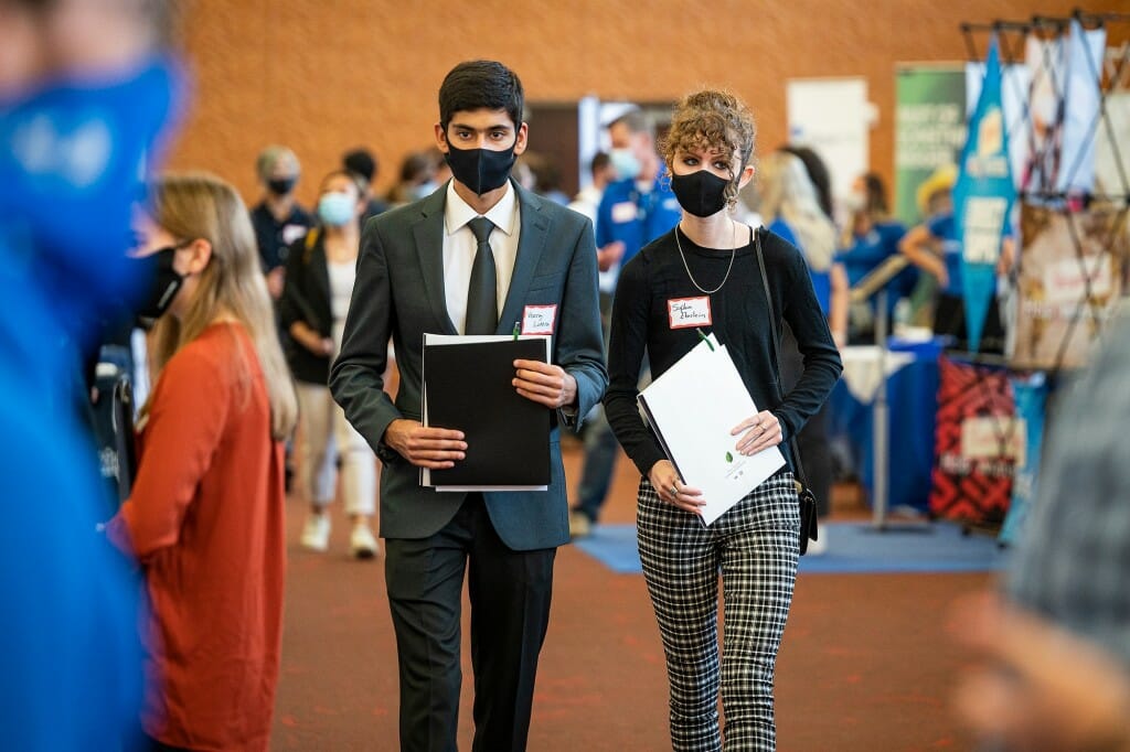 A male student in a suit and tie and a female student in slacks and a sweater walk together holding papers in their hands