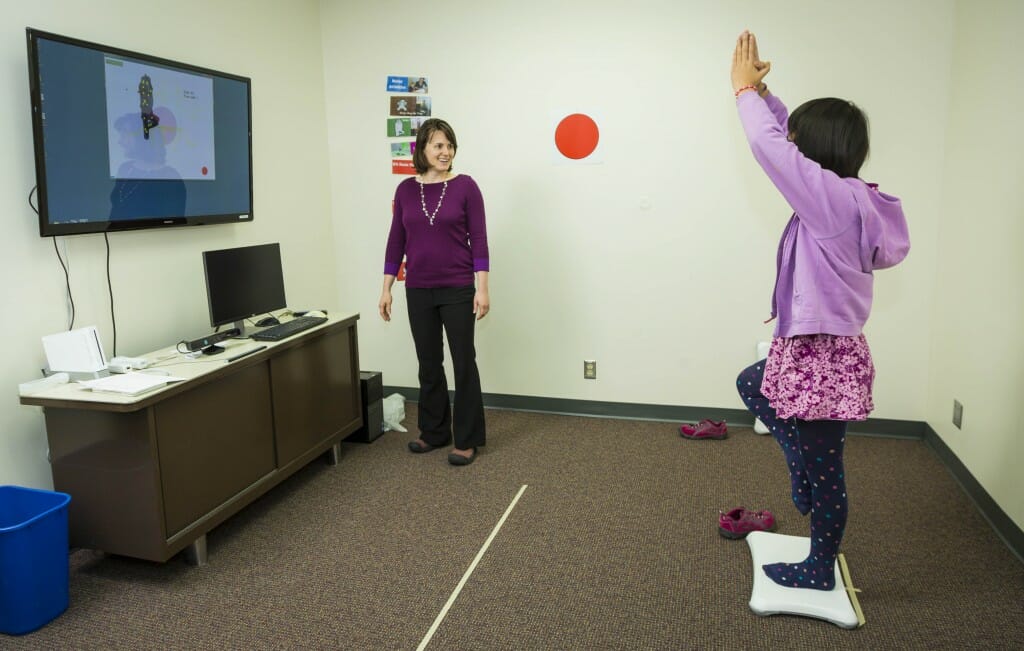 A young person standing on a Wii board while an adult watches next to a video screen