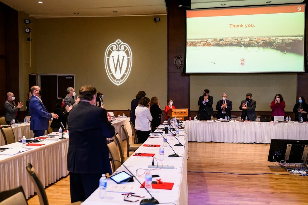 Photo: Board members stand at a table and applaud the chancellor, who stands in front of them.