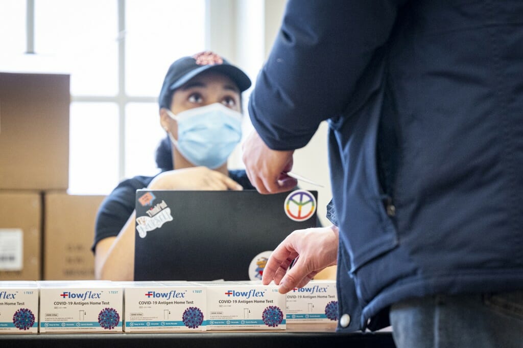 Person wearing face covering sitting at table across from person picking up testing kit