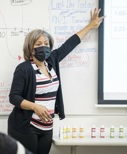 A person with a face covering pointing to a whiteboard in a classroom