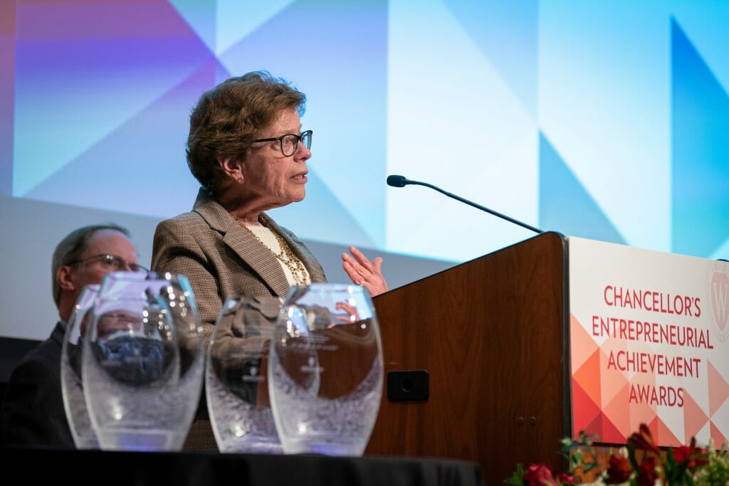 Blank speaking into microphone at podium with glass trophies next to it on a table