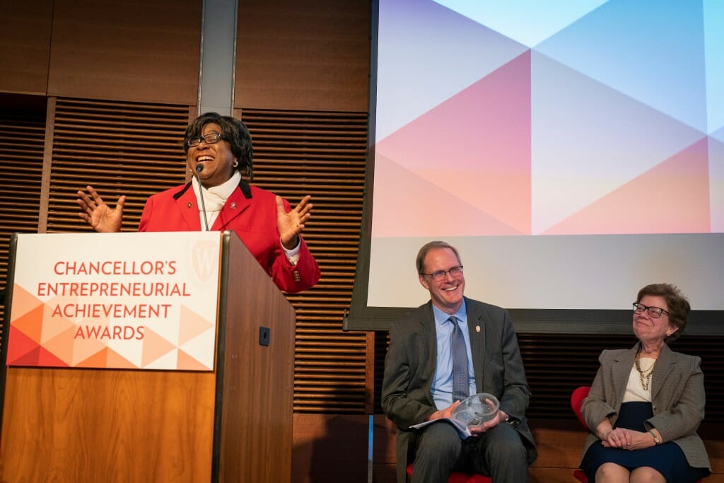 King-Bailey speaking at podium while Blank and unidentified man sit behind her on stage, smiling