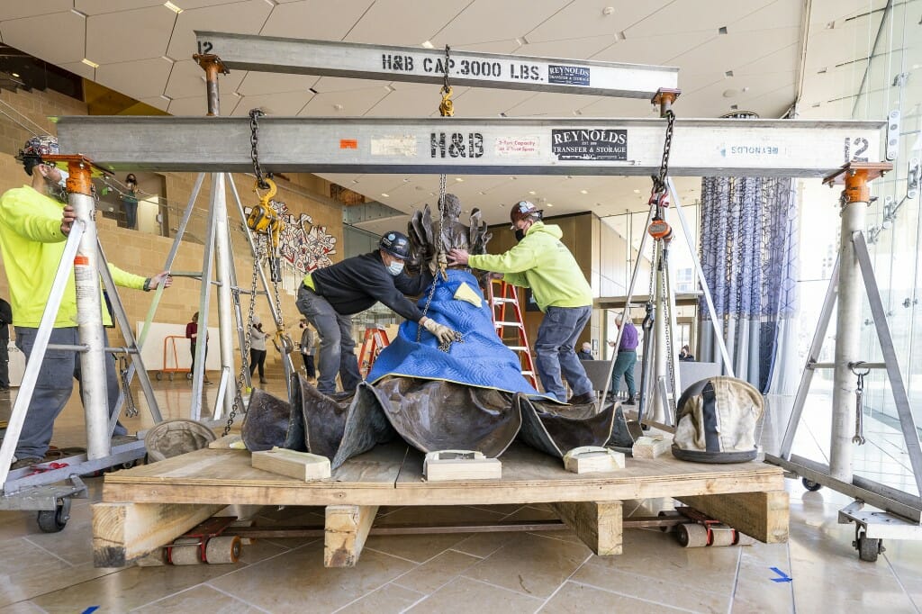 Workers removing protective padding from statue, which is standing on a wooden shipping platform