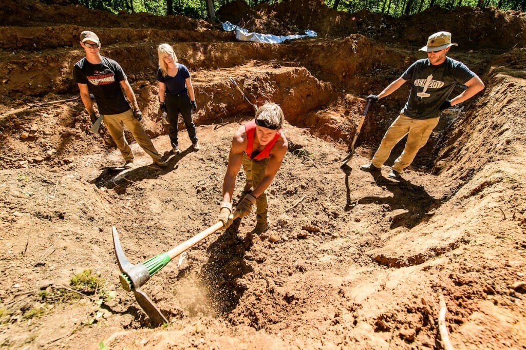 Three people standing in the dirt, one swinging a pickax