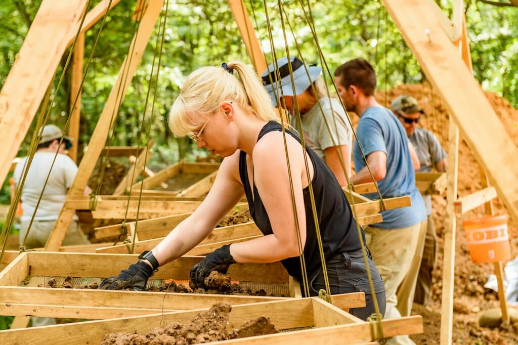 Person reaching into screened wooden box to sift dirt