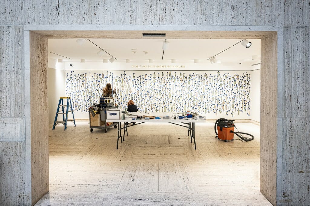 View of large room with 2 people working at table in front of a wall covered in embroidered plants