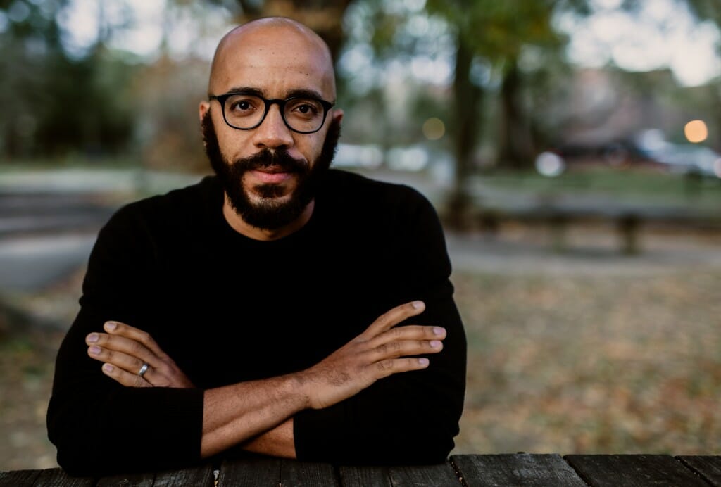 Outdoor portrait of Clint Smith with arms crossed; background is out-of-focus and unidentifiable