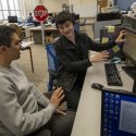 Two people sitting a a desk with one motioning toward a computer monitor