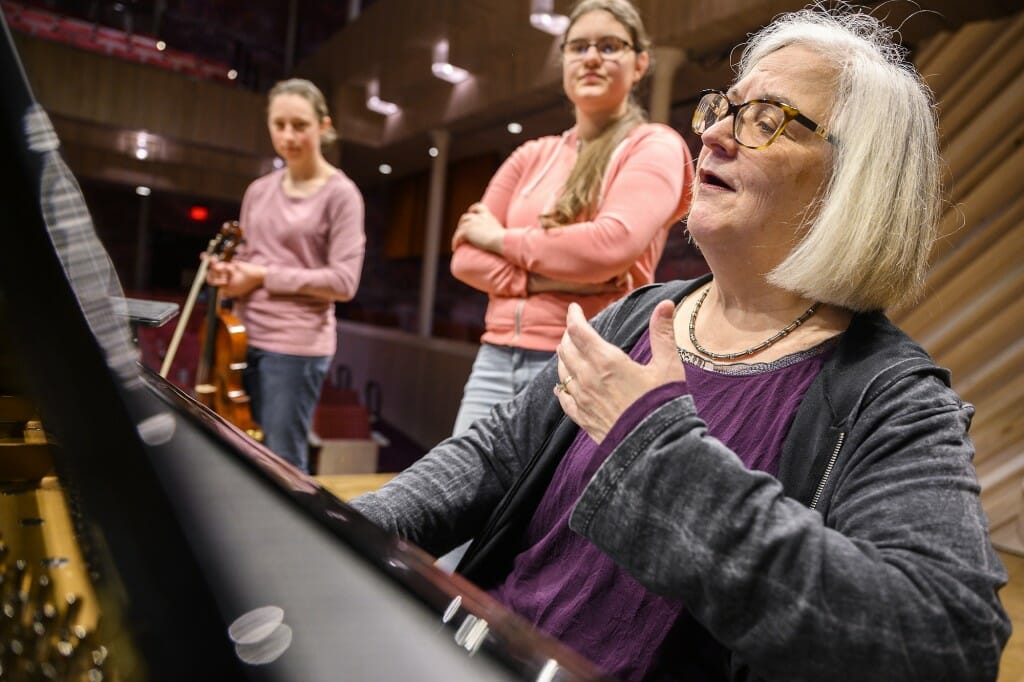 Martha Fischer in a music classroom playing a piano while 2 students stand listening