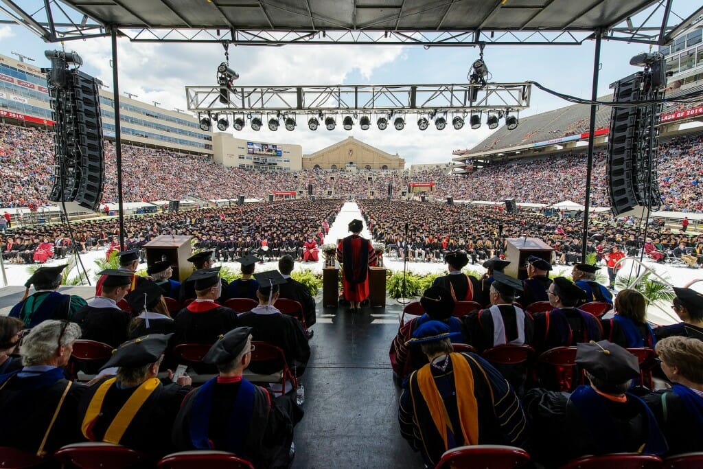 View from back of stage looking toward the bowl of the stadium, with Blank seen from behind addressing students from a podium