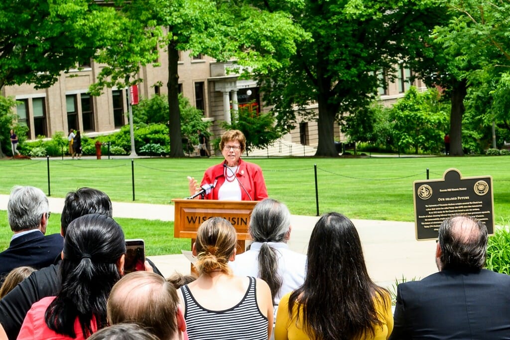 Blank speaking at a podium to a small group seated on Bascom Hill. A plaque is at right, the Education Building is in the background