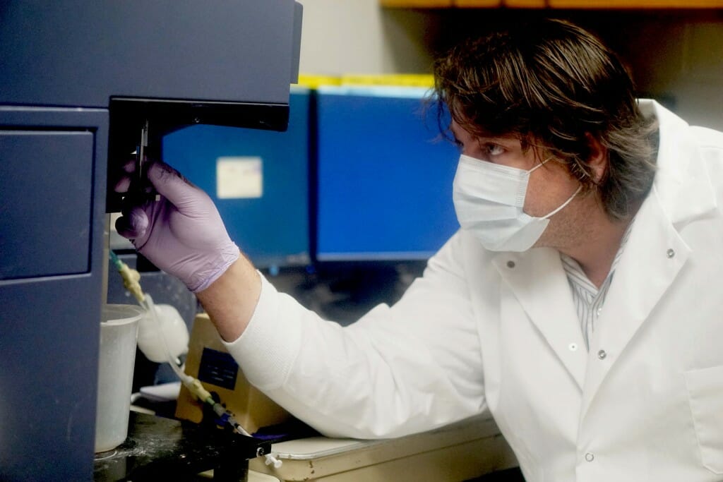 Brock Kingstad-Bakke wearing a surgical grade mask and white lab coat and purple rubber gloves, looking at a lab experiment