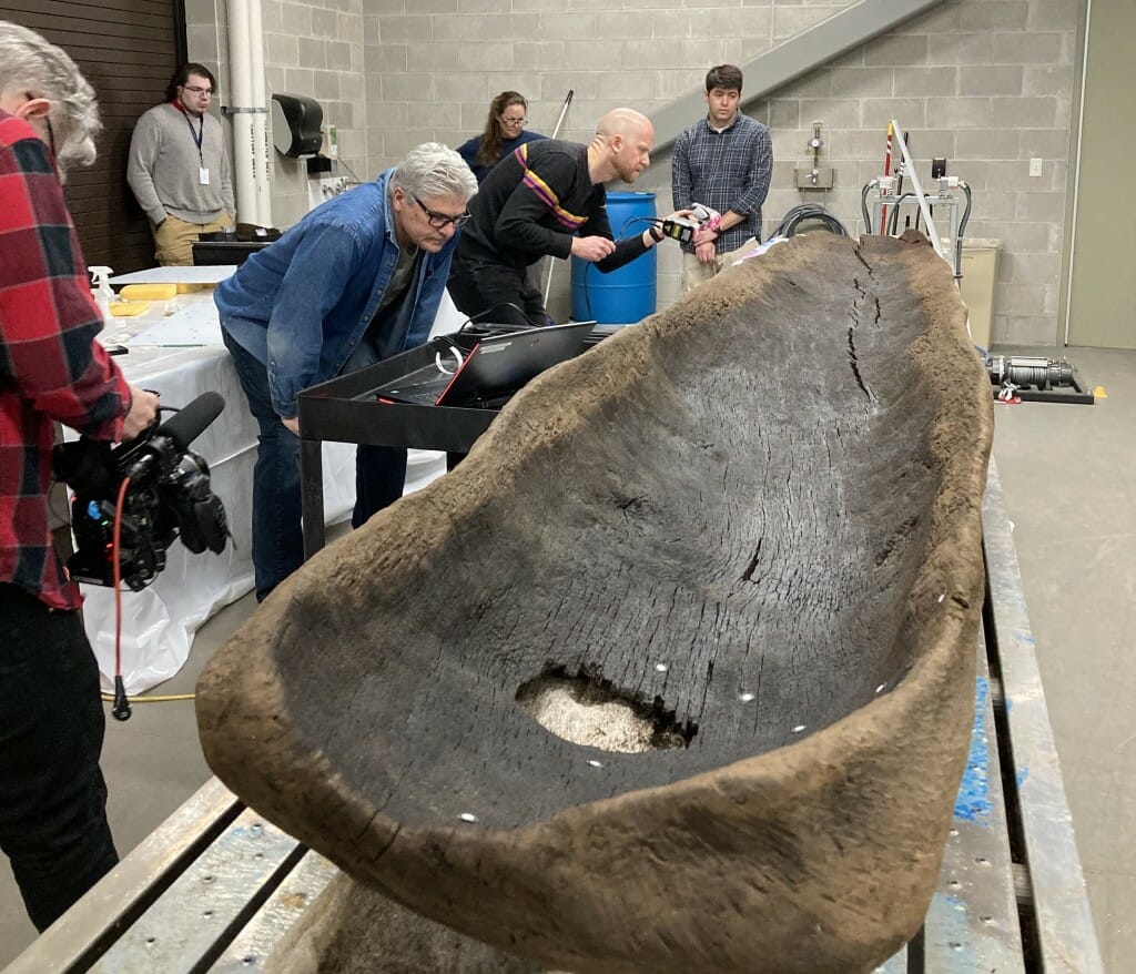 People gather around an old wooden canoe, peering at it through electronic instruments.