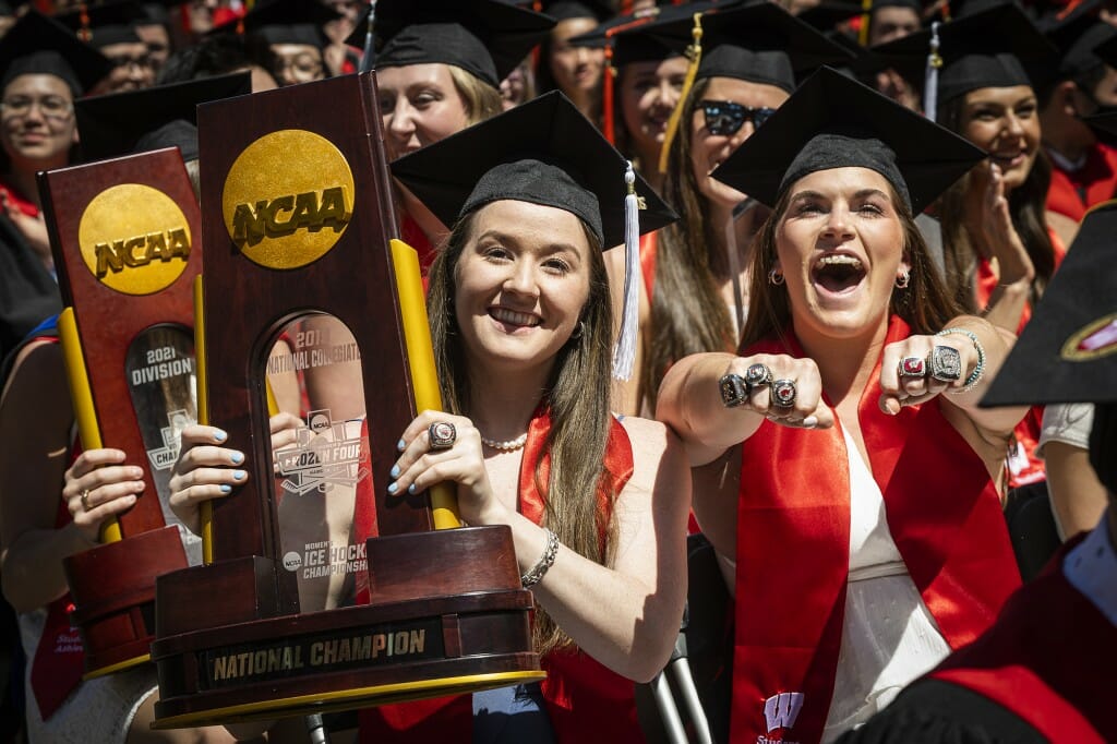 Two graduates holding NCAA championship trophies and showing off championship rings