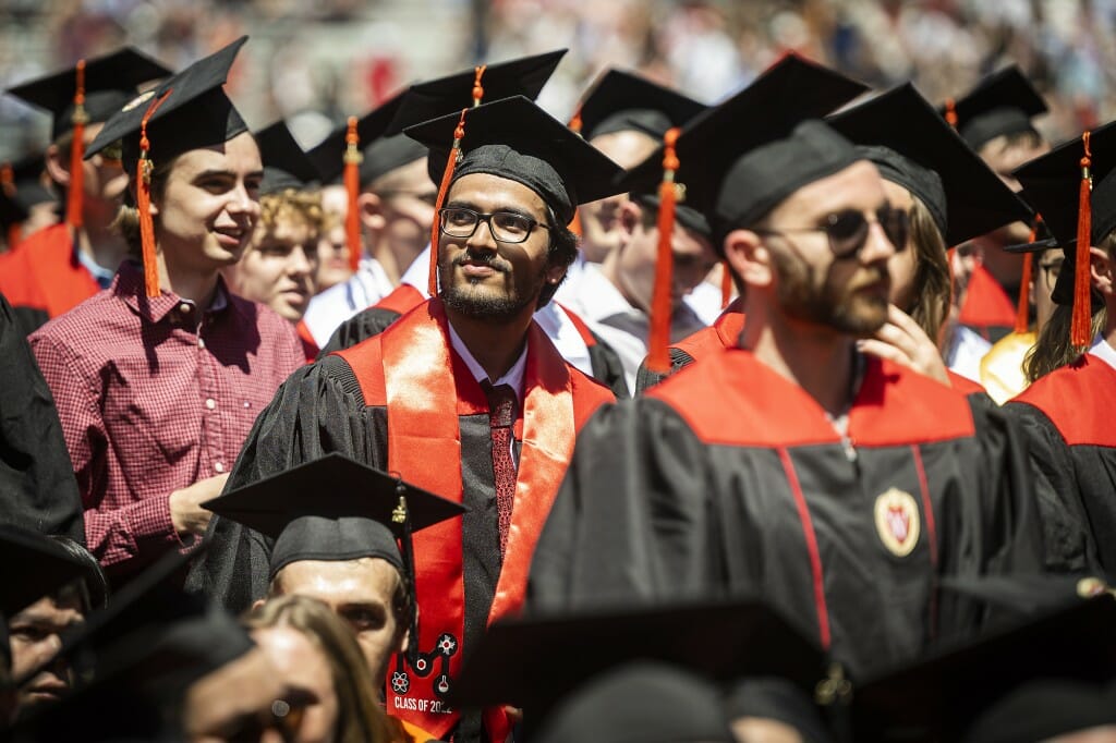 A person in cap and gown standing in the crowd and looking up toward the audience in the bleachers