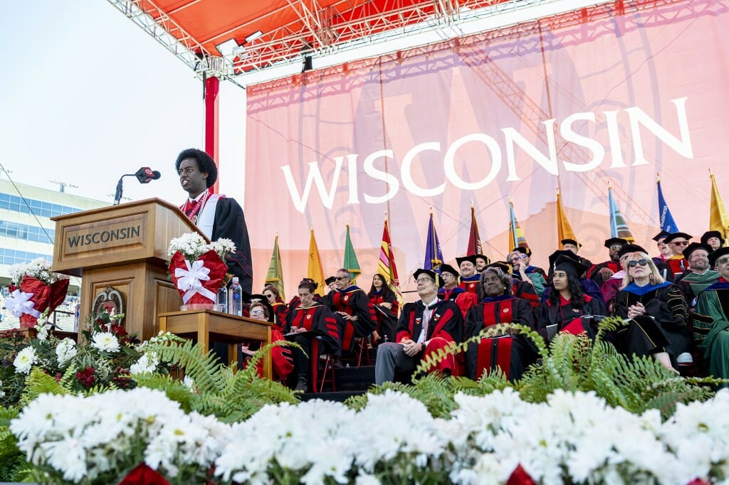 Barni speaking at podium in front of a backdrop reading WISCONSIN in large letters over eight flags