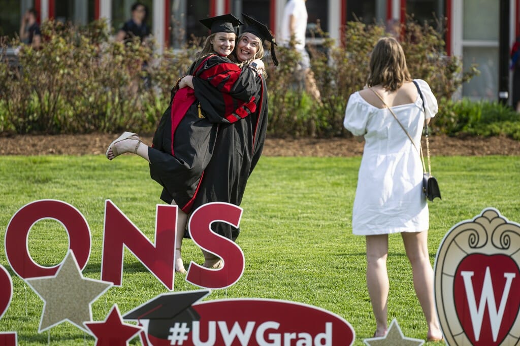 Two people in caps and gowns hugging while a person in a white dress takes their photo on a grassy lawn