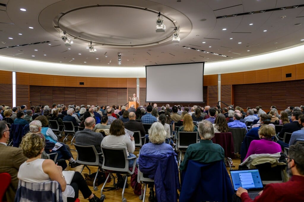 View from the back of the room of large, seated crowd and projection screen above the stage