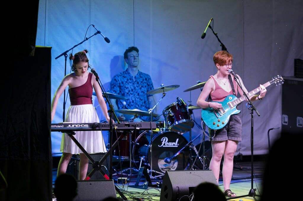 Band members performing on guitar, drums and keyboards on stage