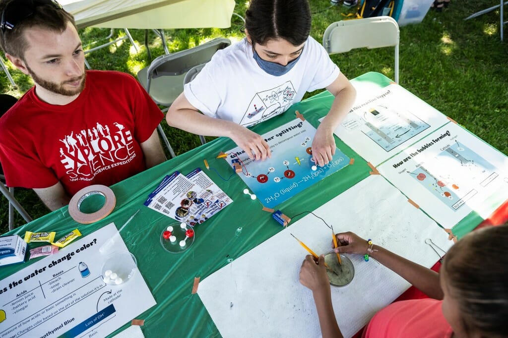 Two college students sitting at a table across from a child touching a scientific instrument